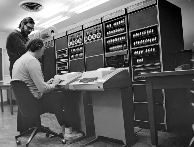 Ken Thompson (sitting) and Dennis Ritchie (standing) in front of a PDP-11. Ritchie annotated this press image for Bell Labs as <a href='https://www.bell-labs.com/usr/dmr/www/picture.html'>"an amusing photo,"</a> and he joked that he had much "more luxuriant and darker hair" at the time of the photo than when it appeared in magazines like the March 1999 Scientific American (which, unfortunately, incorrectly swapped IDs for the two). 